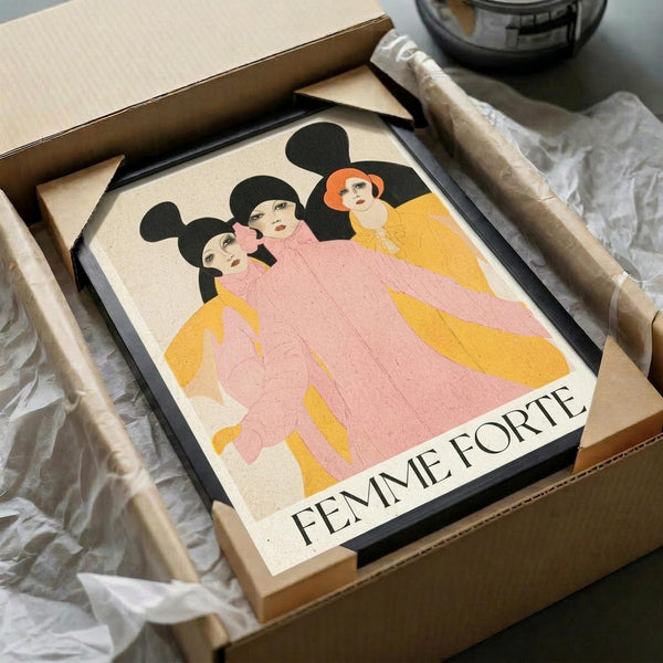 May include: Framed vintage-style poster print featuring three women with black hair in flowing pink and yellow robes or dresses, with "FEMME FORTE" text at bottom. Print has cream background with minimalist illustration style. Black frame sits in opened cardboard shipping box with white tissue paper, alongside rolled tube packaging on white fabric surface.