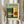 May include: Hands holding a framed Bauhaus architectural poster featuring geometric modernist buildings in green, orange, and cream colors against a sage green background. The vintage-style print shows angular structures with grid windows and bold color blocks. Person wears cream clothing and holds dried pampas grass, creating a styled scene against white curtains and neutral walls.