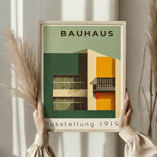 May include: Hands holding a framed Bauhaus architectural poster featuring geometric modernist buildings in green, orange, and cream colors against a sage green background. The vintage-style print shows angular structures with grid windows and bold color blocks. Person wears cream clothing and holds dried pampas grass, creating a styled scene against white curtains and neutral walls.