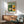 May include: Framed Bauhaus poster with geometric modernist building illustration in green, yellow, and cream colors hanging above wooden credenza with lamp and mid-century green upholstered chair in sunlit room with white walls
