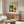 May include: Framed Bauhaus poster with green background featuring modernist architectural building in yellow, orange and cream colors, displayed above wooden mid-century credenza in minimalist living room with white curved chair, round glass coffee table, and floor lamp