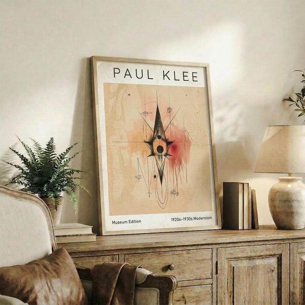 May include: Framed Paul Klee modernist art print displayed on rustic wooden sideboard in warm-toned living room. Abstract artwork features geometric shapes with coral and peach watercolor washes on cream background. Scene includes fern plant, ceramic vase, stacked books, and brown leather furniture in soft natural lighting.