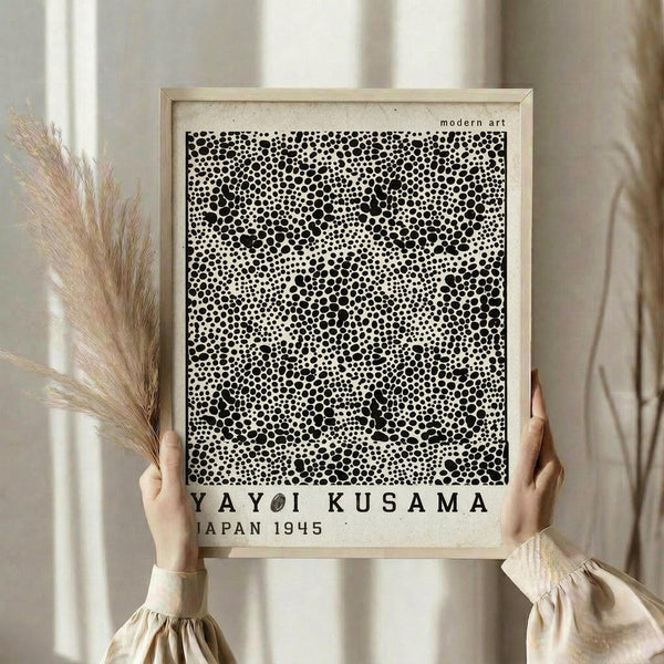 May include: Hands holding a framed black and white polka dot art print in light wooden frame. The artwork features densely packed black circles of varying sizes on white background. Person wears cream ruffled sleeves, holds dried pampas grass, standing against neutral curtained window backdrop in soft natural lighting.