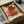 May include: Framed Paul Klee abstract art print in dark frame, featuring geometric rectangular shapes in warm earth tones of red, orange, brown, and yellow on cream background. Print sits in open cardboard box with white tissue paper, alongside rolled paper tube on white fabric surface.