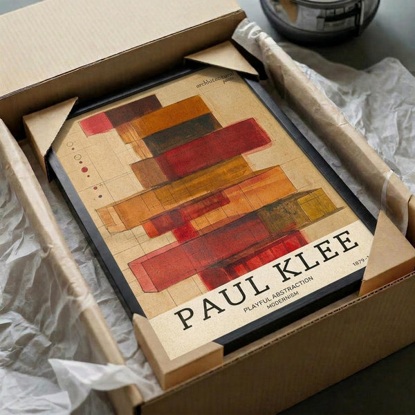May include: Framed Paul Klee abstract art print in dark frame, featuring geometric rectangular shapes in warm earth tones of red, orange, brown, and yellow on cream background. Print sits in open cardboard box with white tissue paper, alongside rolled paper tube on white fabric surface.