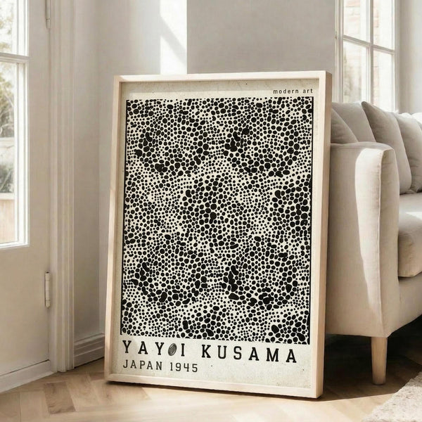 May include: Large framed art print leaning against white wall next to beige sofa in bright living room. Black and white dotted pattern artwork resembles organic cellular or spotted texture with "YAYOI KUSAMA JAPAN 1945" text at bottom. Natural wood frame, approximately 3 feet tall, positioned on light hardwood floor near windows with natural daylight streaming in.