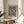 May include: Framed art print with black polka dot pattern on cream background, displayed on round wooden side table next to beige upholstered chair. Natural lighting creates soft shadows on neutral wall. Green plant leaves visible in corner. The artwork features dense, varied-sized dots creating an abstract spotted texture in a light wood frame.
