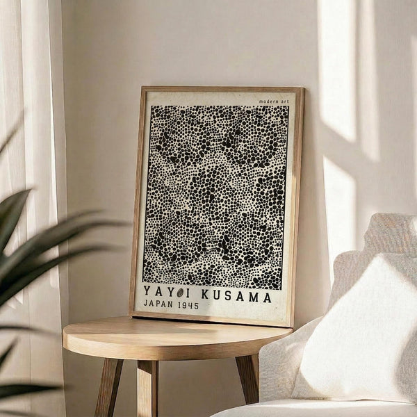 May include: Framed art print with black polka dot pattern on cream background, displayed on round wooden side table next to beige upholstered chair. Natural lighting creates soft shadows on neutral wall. Green plant leaves visible in corner. The artwork features dense, varied-sized dots creating an abstract spotted texture in a light wood frame.