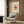 May include: Framed Paul Klee modernist art print hanging on wood-paneled wall above cream upholstered armchair with geometric patterned pillow. Abstract artwork features warm earth tones with black angular shapes and red accent. Modern reading nook with brass floor lamp, dark round side table with open book, and sheer curtains filtering natural light.