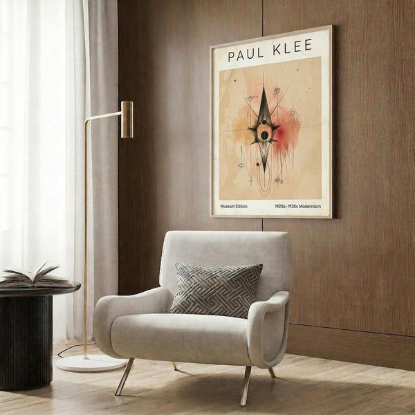 May include: Framed Paul Klee modernist art print hanging on wood-paneled wall above cream upholstered armchair with geometric patterned pillow. Abstract artwork features warm earth tones with black angular shapes and red accent. Modern reading nook with brass floor lamp, dark round side table with open book, and sheer curtains filtering natural light.