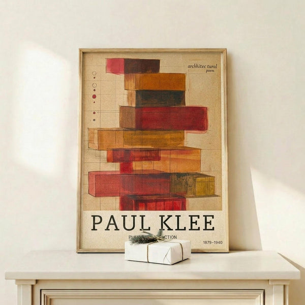 May include: Framed poster displaying abstract geometric composition with stacked translucent rectangular blocks in warm reds, oranges, and earth tones on grid background. Text reads "PAUL KLEE" and "architectural poem" with dates 1879-1940. Light wood frame sits on pale wooden surface beside small white wrapped gift with greenery sprig against cream wall.