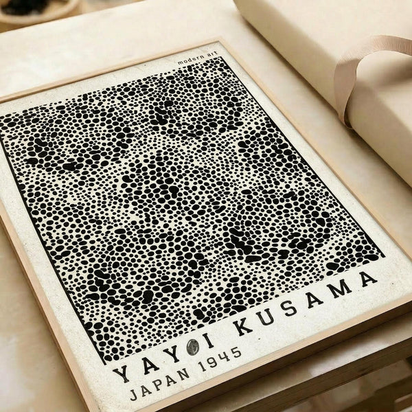 Abstract black and white artwork featuring thousands of varying sized dots creating an organic, spotted pattern reminiscent of cellular structures or bubbles. The piece has "YAYOI KUSAMA JAPAN 1945" text at the bottom and "modern art" at the top. The print is displayed on cream paper with a beige ribbon, photographed against a neutral background with soft lighting that emphasizes the intricate dotted texture and contemporary minimalist aesthetic.