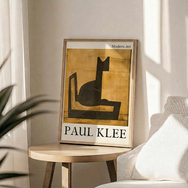 May include: Framed poster featuring Paul Klee's geometric black cat silhouette on golden yellow background, displayed on round wooden side table next to beige upholstered chair. Natural lighting creates soft shadows across cream colored wall. Green plant leaves visible in corner. Modern minimalist interior setting with neutral tones.