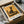 May include: Framed Paul Klee artwork print showing abstract black cat silhouette against warm golden yellow background with geometric lines. Modern art poster displayed in sleek black frame, nestled in cardboard shipping box with protective tissue paper wrapping. Print appears medium-sized with clean white matting, photographed on neutral surface alongside packaging materials and dark glass vessel.