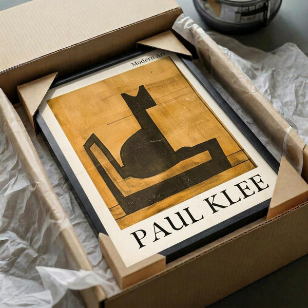 May include: Framed Paul Klee artwork print showing abstract black cat silhouette against warm golden yellow background with geometric lines. Modern art poster displayed in sleek black frame, nestled in cardboard shipping box with protective tissue paper wrapping. Print appears medium-sized with clean white matting, photographed on neutral surface alongside packaging materials and dark glass vessel.