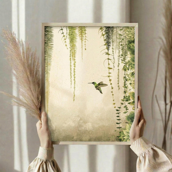 May include: Person in cream ruffled sleeves holding framed botanical wall art featuring green hummingbird flying among hanging ferns and trailing vines on textured beige background, displayed in light wood frame near dried pampas grass against white curtained window