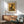 May include: Framed Paul Klee poster featuring abstract black cat silhouette on golden yellow background, displayed above mid-century modern wooden credenza with table lamp. Sage green upholstered armchair with wooden frame sits nearby on wooden floor. Warm natural lighting creates shadows across white wall in modern living room setting.