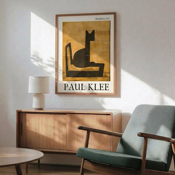 May include: Framed Paul Klee poster featuring abstract black cat silhouette on golden yellow background, displayed above mid-century modern wooden credenza with table lamp. Sage green upholstered armchair with wooden frame sits nearby on wooden floor. Warm natural lighting creates shadows across white wall in modern living room setting.