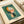 May include: Abstract geometric art print by Wassily Kandinsky displayed on cream textured paper against white fabric. The artwork features overlapping shapes in orange, white, pink, and dark colors against a green-blue cloudy background. Text shows "WASSILY KANDINSKY" at top and "Empor (Upward) Bauhaus Dessau 1924" at bottom. The print appears vintage-style with speckled paper texture.