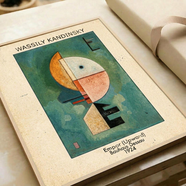 May include: Abstract geometric art print by Wassily Kandinsky displayed on cream textured paper against white fabric. The artwork features overlapping shapes in orange, white, pink, and dark colors against a green-blue cloudy background. Text shows "WASSILY KANDINSKY" at top and "Empor (Upward) Bauhaus Dessau 1924" at bottom. The print appears vintage-style with speckled paper texture.