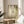 May include: Framed botanical art print featuring green hummingbird flying among cascading fern fronds and hanging vines on cream background, displayed on round wooden side table next to beige upholstered chair in sunlit room with white walls and houseplant