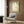 May include: Botanical wall art featuring green hanging ferns and a small hummingbird against a soft cream background, displayed in a gold frame on wood paneled wall above beige upholstered armchair with geometric patterned pillow, alongside brass floor lamp and dark side table with open book