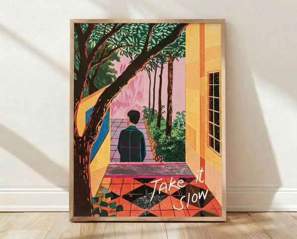 May include: Framed colorful poster print leaning against white wall on wooden floor. Artwork shows person in dark clothing walking on pink checkered path toward building with yellow walls and windows. Large tree with green foliage dominates left side, geometric patterned ground below. White cursive text "Take it Slow" overlays bottom right corner. Natural wood frame, soft shadows cast on wall.