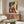 May include: Colorful contemporary art print displayed above wooden credenza in modern living room. Artwork features figure walking through tropical courtyard with palm trees, pink and orange buildings, red tile floor. Room shows white curved chair, glass coffee table, brass floor lamp, and neutral walls with natural lighting.
