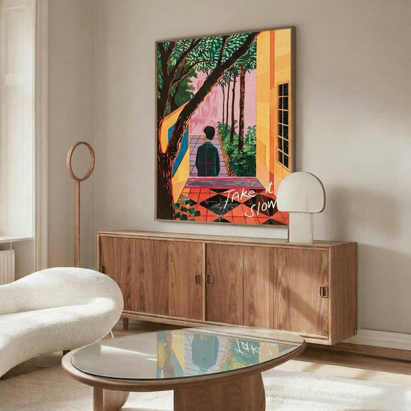 May include: Colorful contemporary art print displayed above wooden credenza in modern living room. Artwork features figure walking through tropical courtyard with palm trees, pink and orange buildings, red tile floor. Room shows white curved chair, glass coffee table, brass floor lamp, and neutral walls with natural lighting.