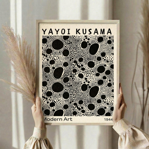 May include: Person in beige ruffle-sleeve top holding framed black and white polka dot art print while standing near dried pampas grass against soft white curtained background. The artwork features various sized black circles and dots in Kusama's signature infinity net style, displayed in light wood frame.