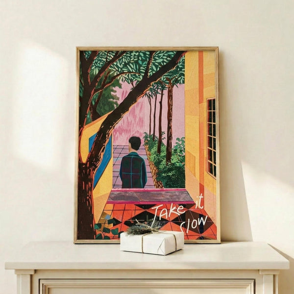 May include: Framed poster displaying a colorful illustration of a person in dark clothing walking through an architectural space with yellow walls, large tree with green foliage, pink background elements, and red geometric floor patterns. White text reads "Take it Slow" across the lower portion. The artwork is displayed in a thin wooden frame on a light wood shelf alongside a small white wrapped gift box against a cream wall.