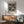 May include: Framed black and white polka dot art print hanging above wooden sideboard in modern living room. Green upholstered chair with wooden arms sits nearby, white table lamp on credenza. Natural sunlight casts shadows on white wall. Print features scattered circles of varying sizes creating abstract dotted pattern in minimalist style.