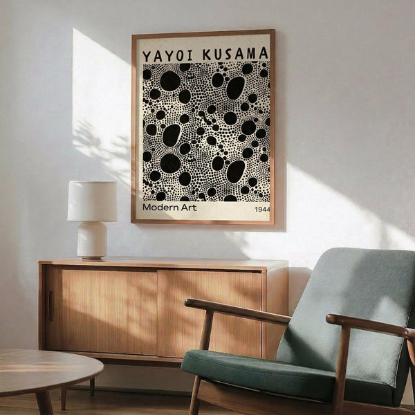 May include: Framed black and white polka dot art print hanging above wooden sideboard in modern living room. Green upholstered chair with wooden arms sits nearby, white table lamp on credenza. Natural sunlight casts shadows on white wall. Print features scattered circles of varying sizes creating abstract dotted pattern in minimalist style.