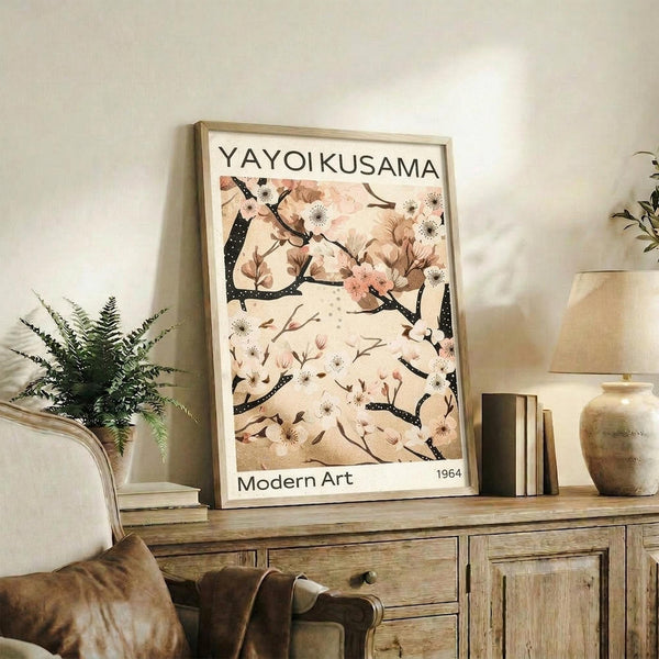 May include: Framed poster print displayed on rustic wood sideboard in neutral-toned living room. Artwork features delicate cherry blossom branches with pink flowers and dark spotted branches on cream background, with "YAYOI KUSAMA" and "Modern Art 1964" text. Scene includes fern plant, ceramic vases, books, and beige upholstered furniture in soft natural lighting.