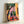 May include: Framed colorful poster print leaning against white wall. Artwork shows person in plaid shirt viewed from behind, standing in vibrant indoor space with large tree, yellow architectural elements, pink walls, and orange tiled floor. White cursive text reads "take it slow" in corner. Modern illustration style with bold colors and geometric patterns.