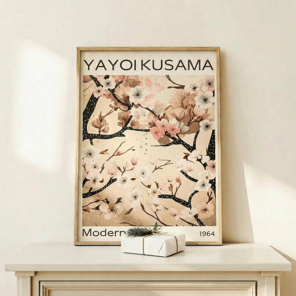 May include: Framed art print featuring delicate cherry blossom branches with pink flowers and dark spotted stems against a cream background, displayed on a light wood shelf next to a small wrapped gift with greenery. The minimalist poster has "YAYOI KUSAMA" at the top and "Modern 1964" at the bottom, housed in a thin natural wood frame against a white wall.