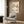 May include: Framed poster featuring Japanese botanical artwork with cherry blossoms and dark branches on cream background, displayed on wood-paneled wall above beige upholstered armchair with geometric patterned pillow. Modern brass floor lamp and dark round side table with open book complete the neutral-toned reading nook setting.