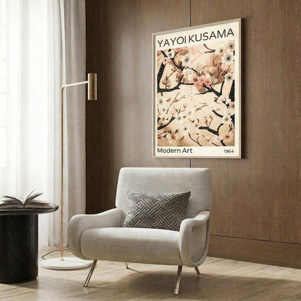 May include: Framed poster featuring Japanese botanical artwork with cherry blossoms and dark branches on cream background, displayed on wood-paneled wall above beige upholstered armchair with geometric patterned pillow. Modern brass floor lamp and dark round side table with open book complete the neutral-toned reading nook setting.