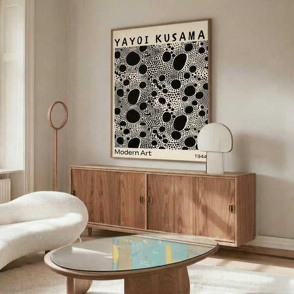 May include: Black and white polka dot art print with "YAYOI KUSAMA" and "Modern Art 1944" text, displayed in thin wooden frame above mid-century walnut credenza. Modern living room setting with cream walls, white upholstered chair, brass floor lamp, oval glass coffee table with colorful abstract surface, and natural lighting from window.