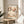 May include: Framed art print displayed on light wood round side table next to beige upholstered chair. Print features delicate cherry blossom branches with pink and white flowers against cream background, black dotted branches, text reading "YAYOI KUSAMA" at top and "Modern Art 1964" at bottom. Natural lighting creates soft shadows on white walls, with plant leaves visible in corner.