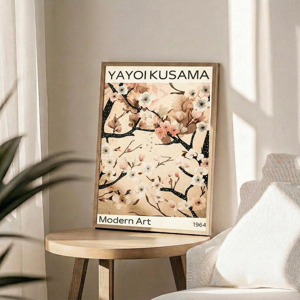 May include: Framed art print displayed on light wood round side table next to beige upholstered chair. Print features delicate cherry blossom branches with pink and white flowers against cream background, black dotted branches, text reading "YAYOI KUSAMA" at top and "Modern Art 1964" at bottom. Natural lighting creates soft shadows on white walls, with plant leaves visible in corner.