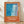 May include: Framed poster with orange background and abstract swimming pool artwork in blues and whites, featuring French text "Plonge dans le grand bain". The print shows wavy water patterns, geometric tiles, and splashing effects. Natural wood frame leaning against white wall on wooden floor, with modern wall sconce visible to the left. Soft natural lighting creates subtle shadows on the wall.