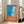 May include: A framed art print with French text "Plonge dans le grand bain" on orange background, featuring abstract blue water textures and geometric patterns in turquoise and navy. The white-framed poster leans against a white wall next to a beige sofa in a bright, modern living room with large windows and natural lighting.