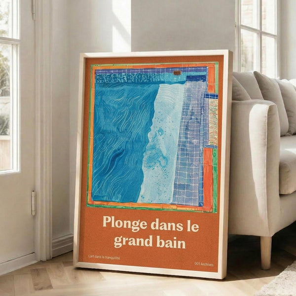 May include: A framed art print with French text "Plonge dans le grand bain" on orange background, featuring abstract blue water textures and geometric patterns in turquoise and navy. The white-framed poster leans against a white wall next to a beige sofa in a bright, modern living room with large windows and natural lighting.