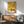 May include: Abstract art poster with yellow and cream background featuring black geometric shapes, colorful dots, and curved lines in Miro's surrealist style. Wooden-framed print hangs above mid-century modern walnut credenza with white ceramic lamp. Sage green upholstered chair with wooden arms sits nearby. Natural sunlight creates shadows on white wall. Indoor plant visible on left side of minimalist living room setting.