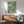 May include: Framed vintage-style swimming poster with turquoise water and three divers in red swimwear, displayed above a wood credenza with white ceramic lamp in modern living room. Mid-century teal armchair and round coffee table visible. Natural sunlight creates geometric shadows on white wall.