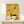 May include: Framed abstract art print in light wood frame leaning against white wall on pale wooden surface. Yellow and cream background features black geometric shapes, curved lines, crescent moon, circles, and organic forms in Miró's distinctive surrealist style. Small white wrapped gift with greenery sits in front of frame, showing scale of approximately 16x20 inch artwork in minimalist room setting.