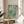 May include: Vintage-style poster with turquoise watercolor background showing three swimmers diving underwater. Features distressed, weathered texture with cream and white areas. Red text reads "DEEP DIVE" with smaller subtitle below. Displayed in thin wooden frame on round wooden side table next to beige upholstered chair with white pillow. Natural lighting creates soft shadows, with plant leaves visible in corner of bright, minimalist room setting.