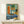 May include: Framed mid-century modern style poster with geometric shapes in blue, orange, and green tones featuring a vinyl record turntable scene. French text reads "Douce nuit sous les étoiles" at bottom. Artwork displayed on light wood shelf next to small wrapped white gift box with natural twine and dried sprig against cream wall.
