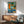 May include: Abstract vintage-style poster featuring turquoise record player with geometric shapes in orange, green, and blue tones, displayed above mid-century modern wooden credenza with table lamp, alongside teal upholstered armchair in minimalist white-walled room with natural lighting